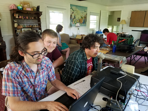 People sit at a desk in front of ham radio equipment as they participate in a radiosport event