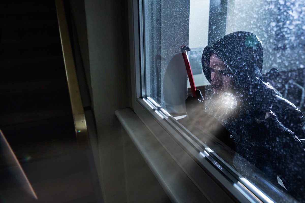 white male thief holding a crowbar and wearing a hood peering out the window of the house